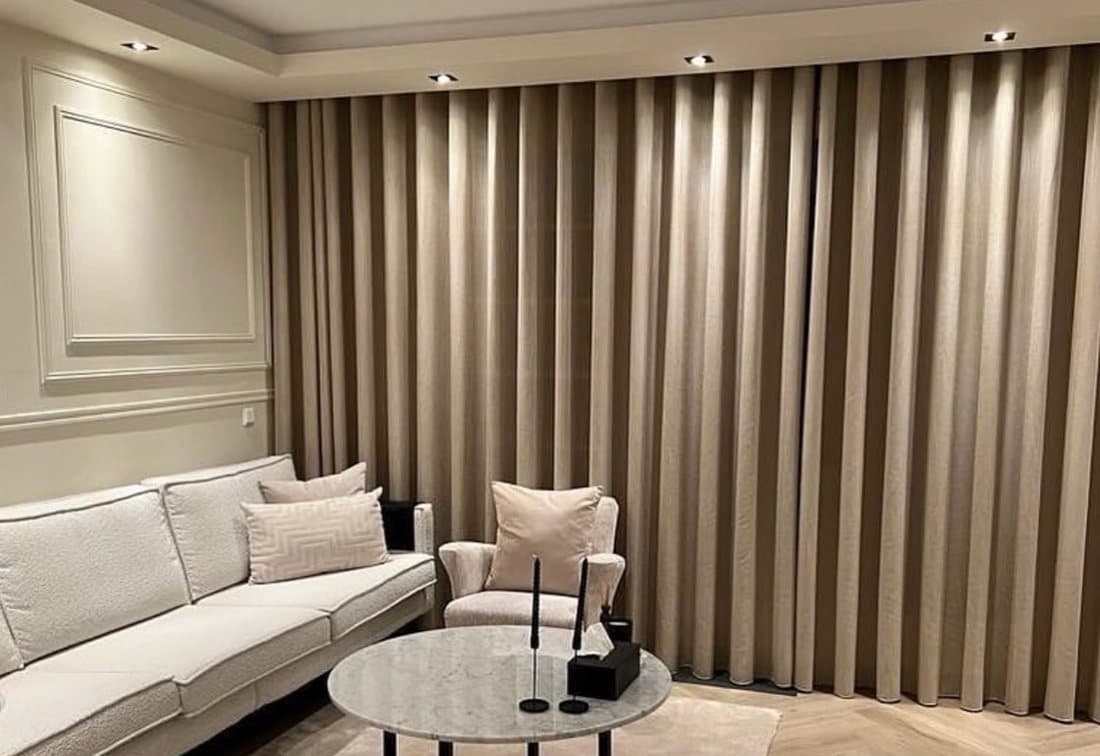 Zitkamer met een wand vol taupe wave-overgordijnen, aangelicht door spots