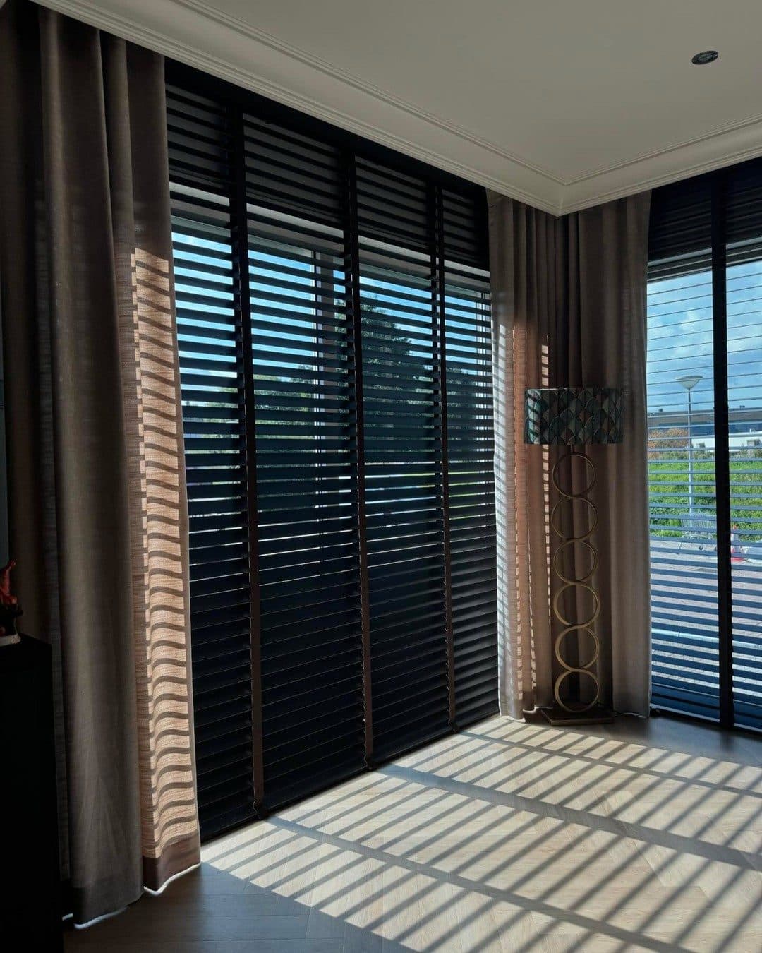 Woonkamer met houten jaloezieën en overgordijnen; late middagzon legt lange strepen over de vloer