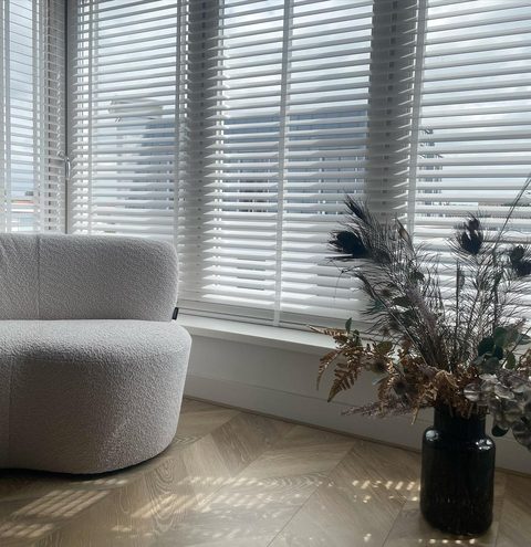 Witte houten jaloezieën over een brede raampartij, met een bouclé fauteuil en een zwarte vaas met droogbloemen
