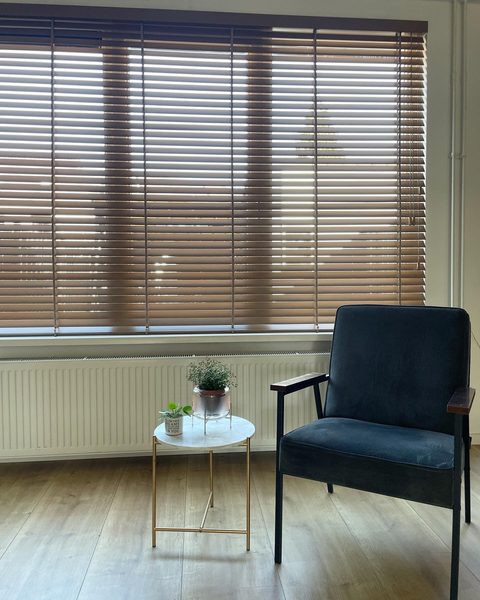 Breed raam met houten jaloezie in warme houtkleur, daaronder een radiator, bijzettafeltje met plantjes en een donkere fauteuil