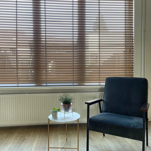 Vierkante uitsnede van een breed raam met houten jaloezie, radiator, marmeren bijzettafeltje en donkerblauwe fauteuil