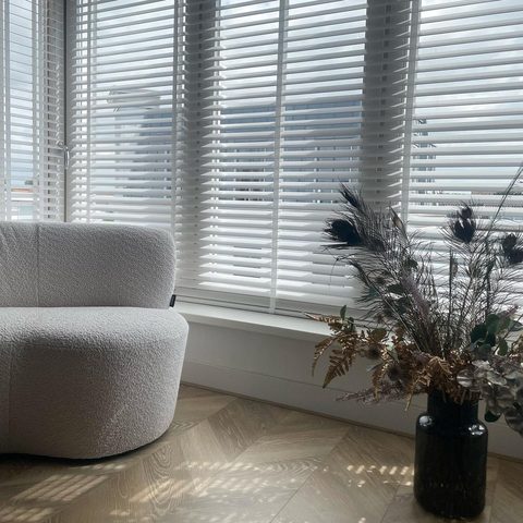 Witte houten jaloezieen met ladderband voor een hoekraam, met een boucle fauteuil en een vaas droogbloemen op visgraatvloer