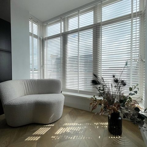 Witte houten jaloezieen langs een brede erker, met een boucle fauteuil, droogboeket en zonlicht op de visgraatvloer