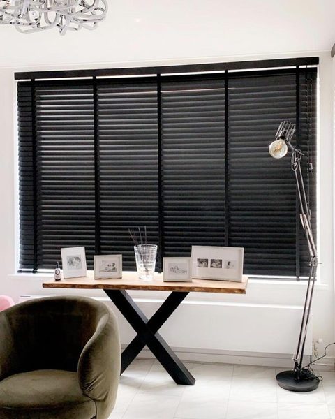Witte kamer met zwarte houten jaloezieen voor een breed raam, een boomstamtafel met fotolijstjes en een groene fauteuil