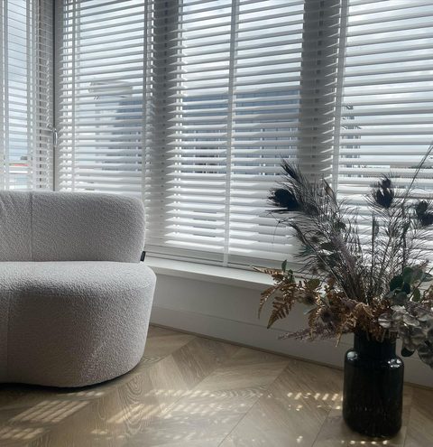 Hoek van een lichte kamer met witte houten jaloezieën, een bouclé fauteuil en een vaas met droogbloemen