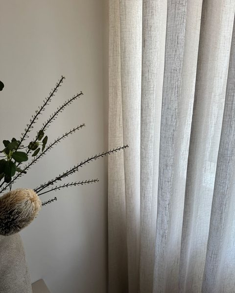 Detail van een linnen inbetween-gordijn in plooien naast een lichte wand met takken in een vaas