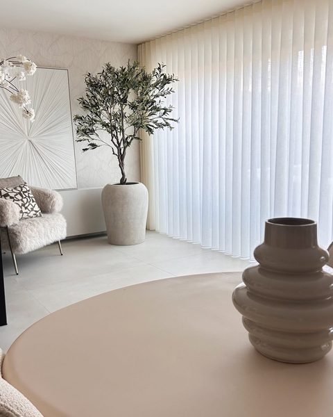 Lichte woonkamer met witte halfdoorschijnende gordijnen in golfplooi over de hele wand, olijfboom en vaas