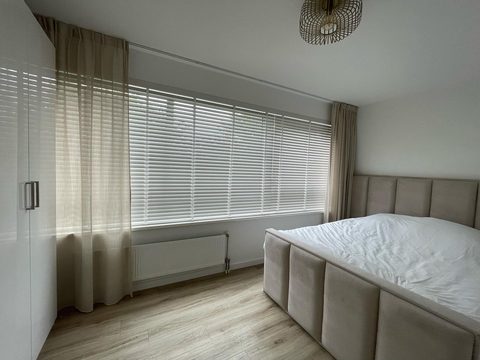 Slaapkamer met witte houten jaloezie voor een breed raam en beige gordijnen aan weerszijden, met boxspring rechts