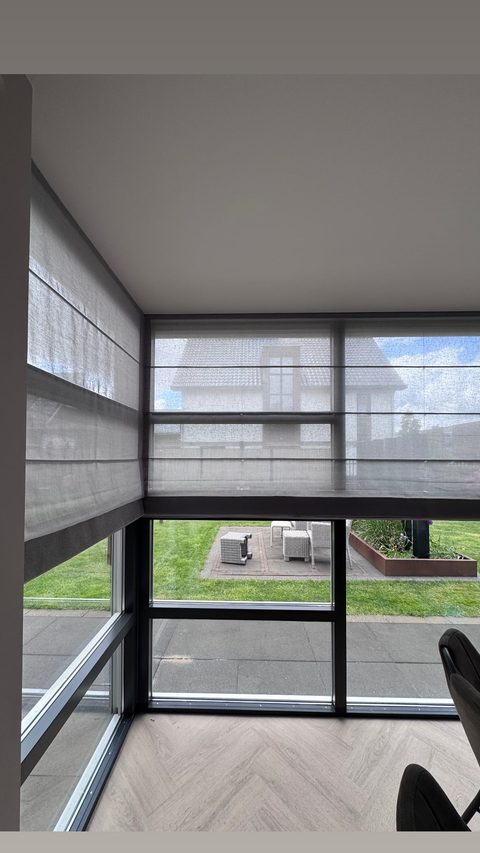Hoekraam met transparante grijze vouwgordijnen van screenstof en uitzicht op de tuin, met een bureaustoel rechtsonder
