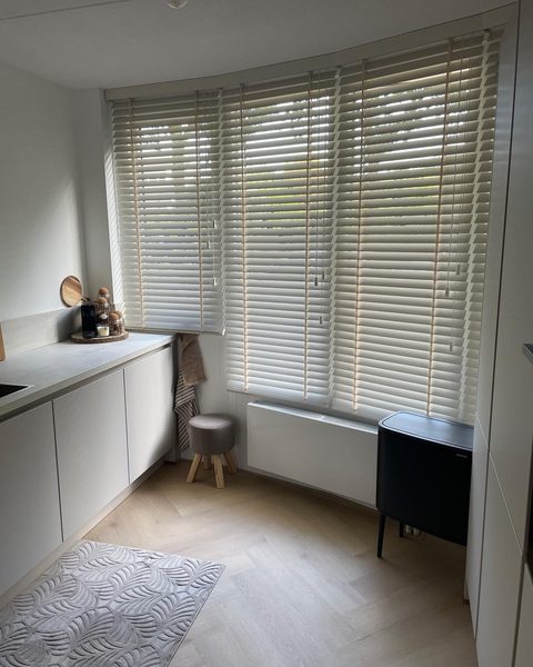 Opgeruimde lichte keukenhoek met witte houten jaloezieen voor een erker en een krukje bij de vensterbank